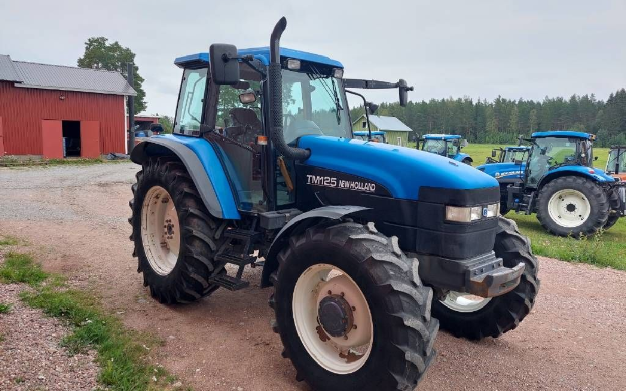 Sininen New Holland -traktori pysäköitynä sorapolulle punaisen navetan lähelle, ja takana pellolla näkyy muita traktoreita.