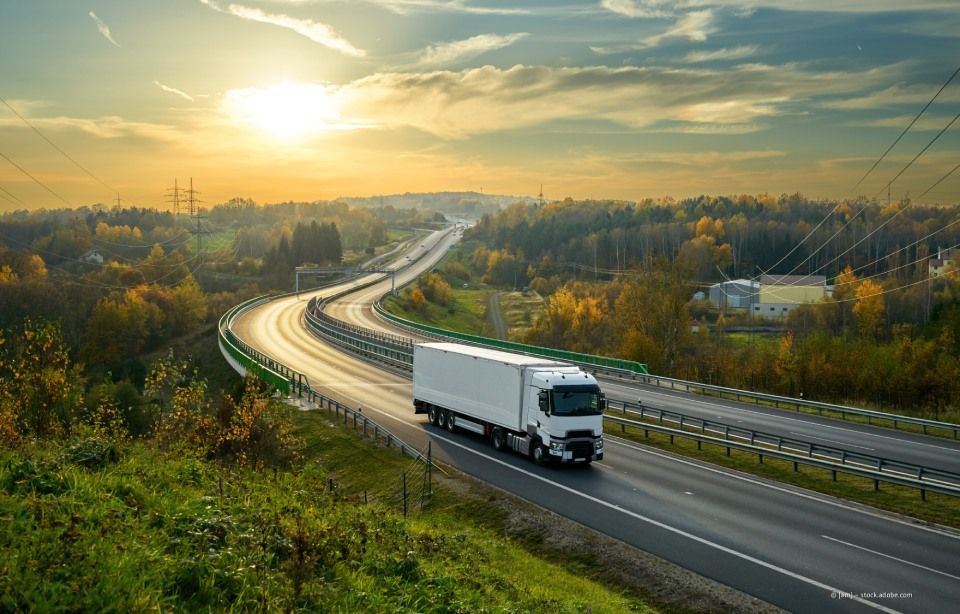 Ein weißer Lastwagen fährt bei Sonnenaufgang auf einer kurvenreichen Autobahn durch eine grüne Landschaft.