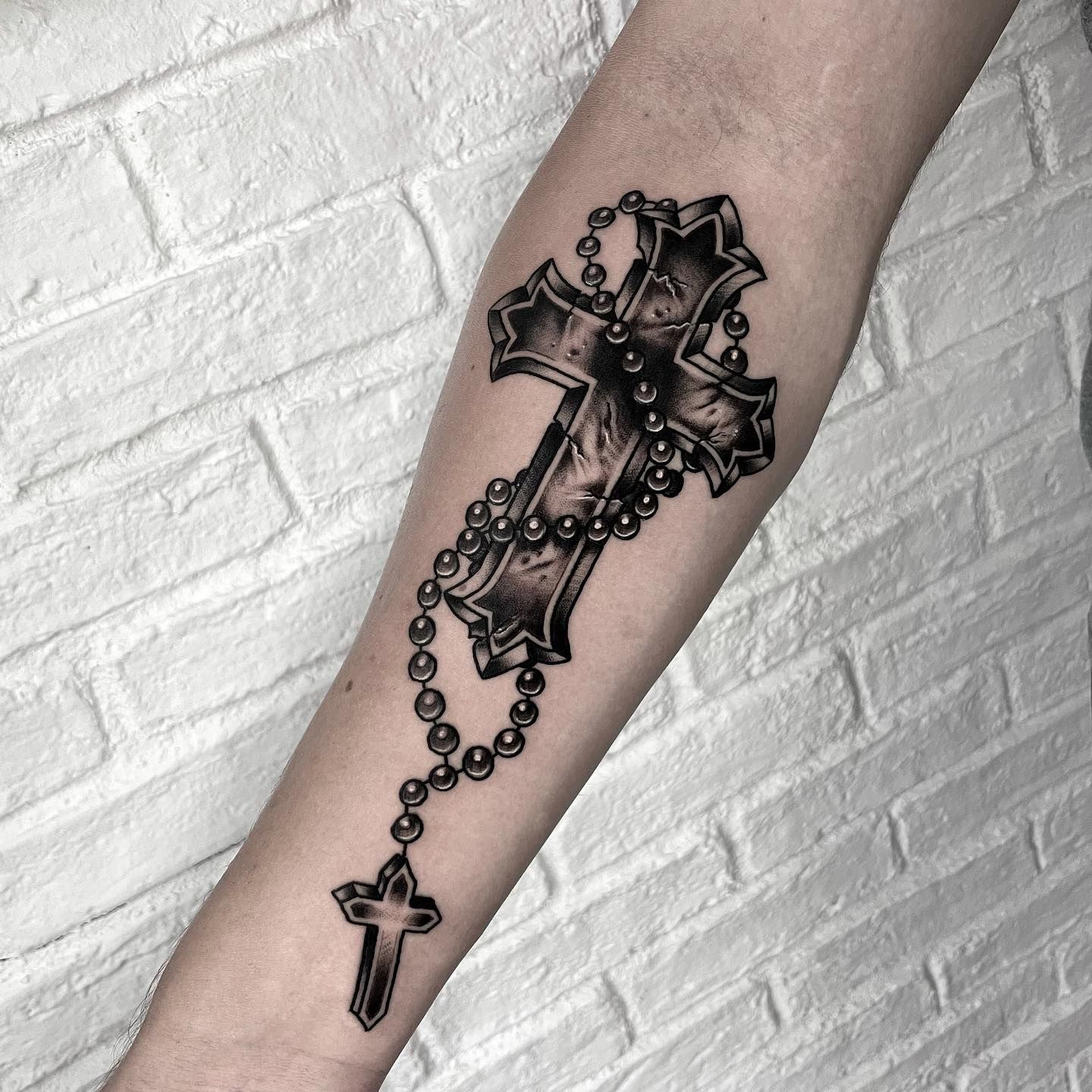 Un tatuaje en blanco y negro de una cruz y un rosario en el antebrazo de una persona.