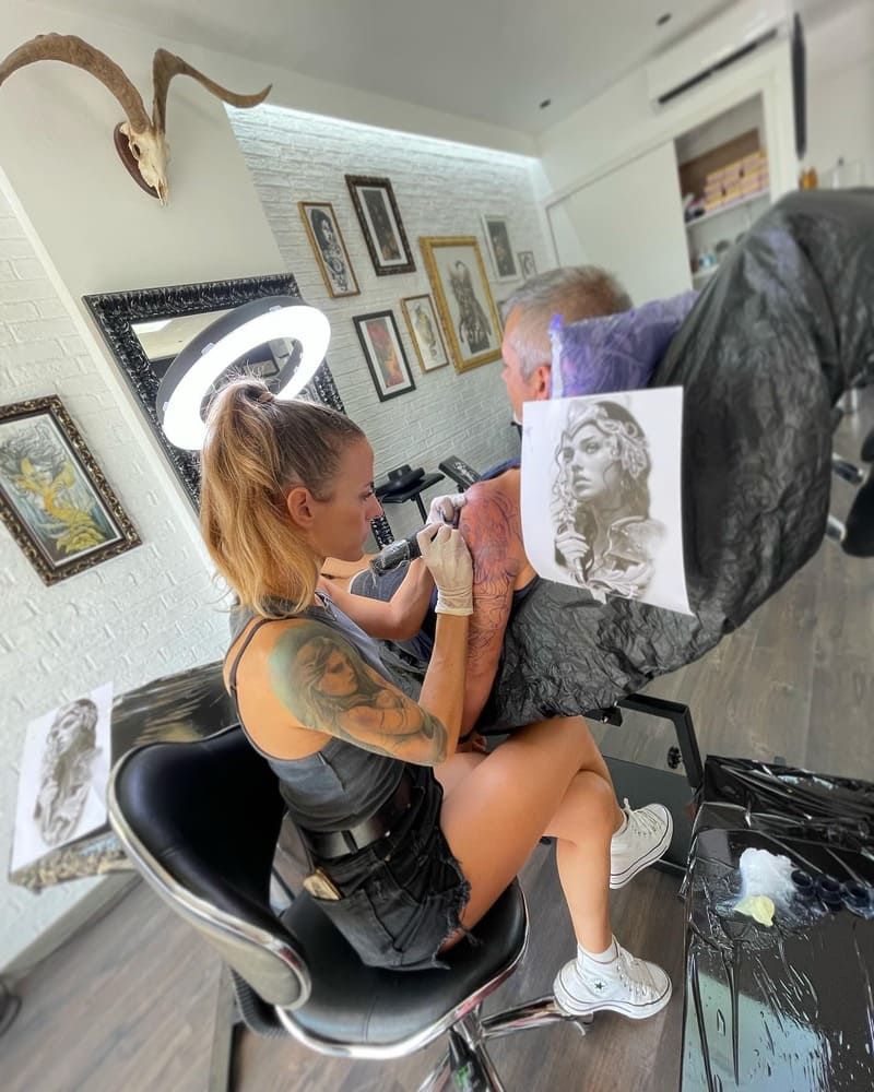 Una mujer está sentada en una silla haciéndose un tatuaje en el brazo de un hombre.