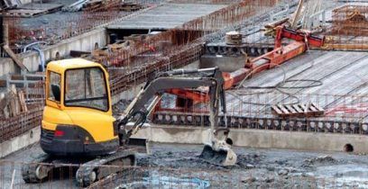 Une pelleteuse jaune sur un chantier de construction en béton, avec des barres d'armature et une longue machine orange en arrière-plan.