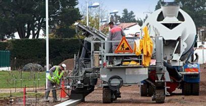 Des ouvriers du bâtiment posent une bordure en béton à l'aide d'une machine ; une bétonnière est visible en arrière-plan.