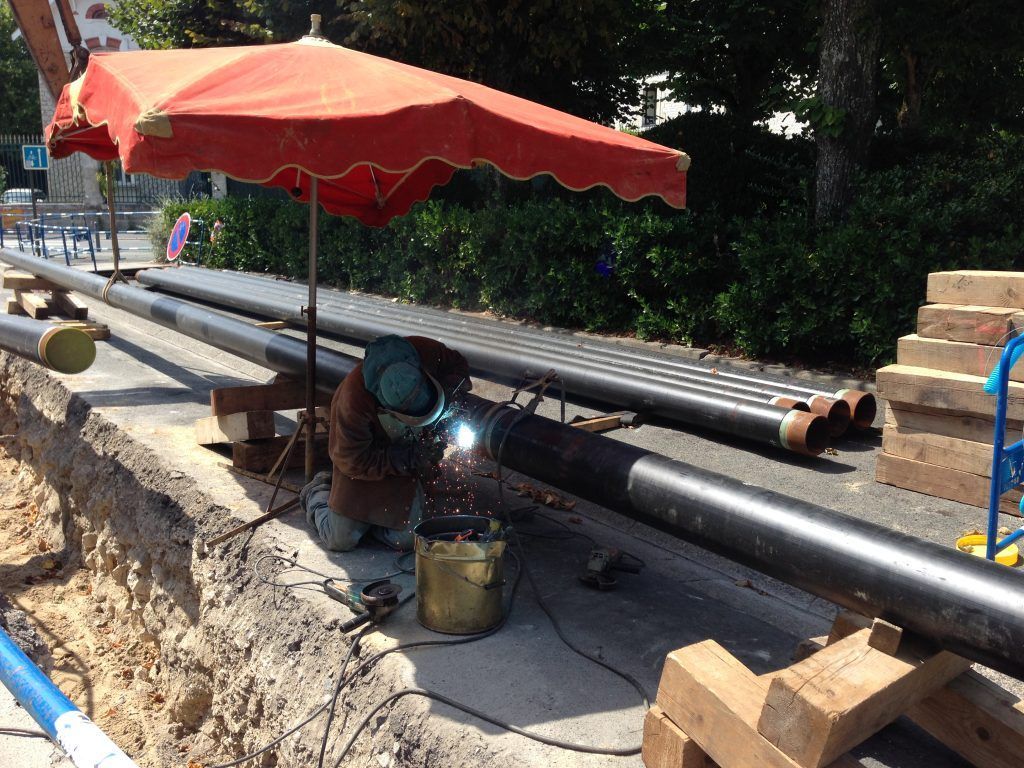 Un soudeur travaille sur de gros tuyaux noirs dans une tranchée, à l'ombre d'un parasol rouge.