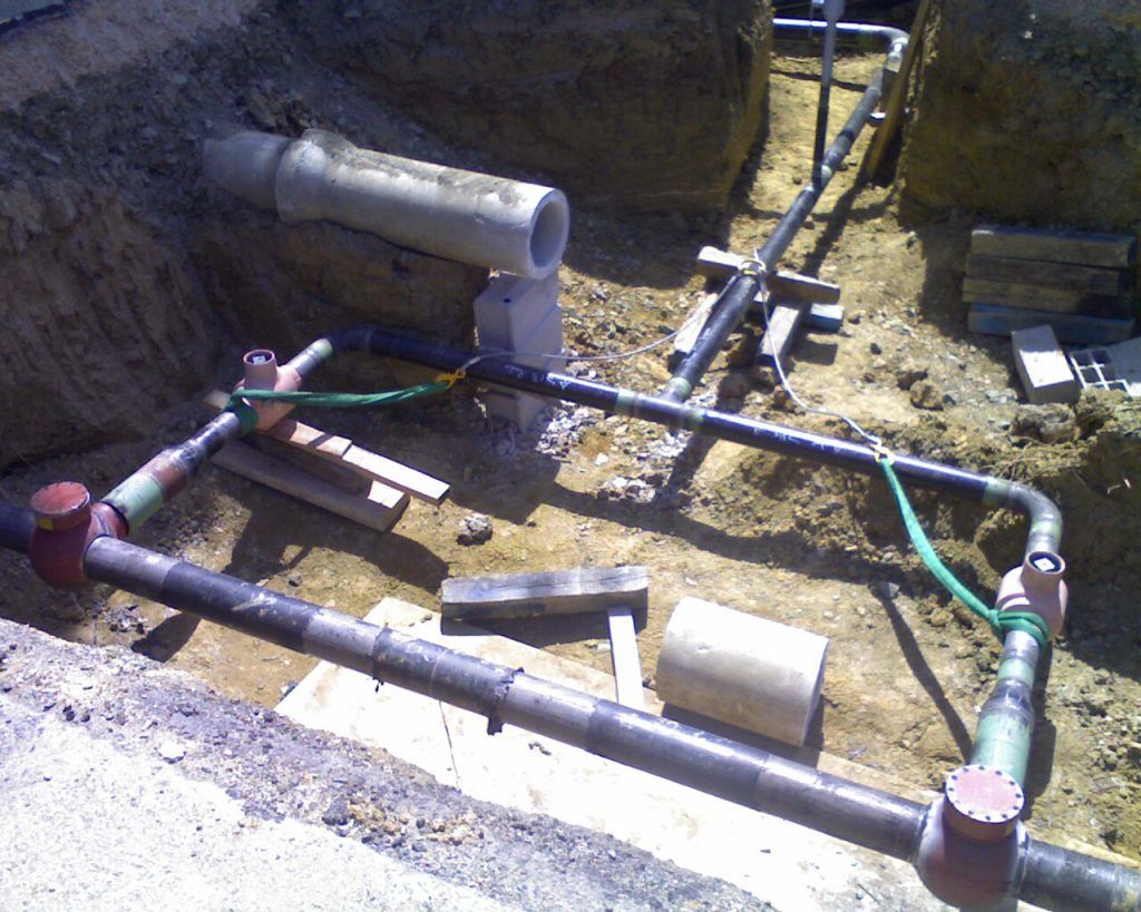 Tuyaux et raccords disposés dans une tranchée, probablement pour la plomberie ou les réseaux. Fils de terre verts visibles.