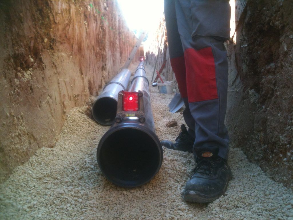 Un ouvrier travaille dans une tranchée, près de gros tuyaux noirs. Un niveau laser rouge est fixé dessus. Le sol est jonché de petits cailloux blancs.