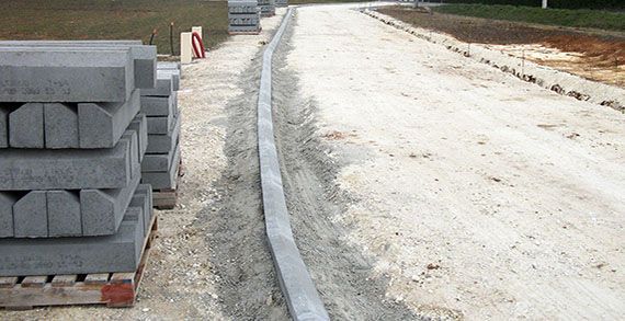 Chantier de construction avec des blocs de bordure en béton empilés et une bordure partiellement installée le long d'un chemin de gravier.