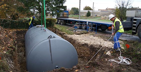 Des ouvriers installent une grande cuve de stockage souterraine grise dans une tranchée, avec un camion plateau en arrière-plan.
