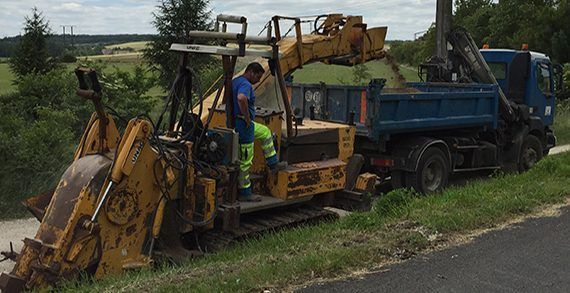 Une fraiseuse routière jaune charge des débris dans un camion-benne bleu. Deux ouvriers en gilets haute visibilité se tiennent à proximité.