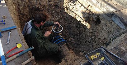 Une personne travaillant sur des canalisations dans une tranchée, tenant un seau et des outils, près d'un trottoir.