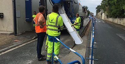 Des ouvriers, portant des gilets de sécurité, déversent du béton depuis un camion dans une rue.