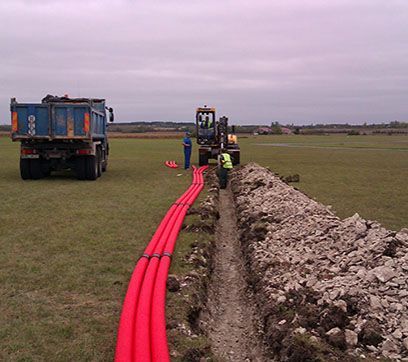 Des ouvriers du bâtiment installent des conduits rouges dans une tranchée au milieu d'un champ, à proximité d'un camion et d'une excavatrice.