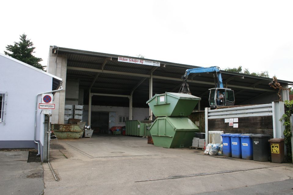 Ein Kran hebt einen grünen Müllcontainer in einem Recyclingzentrum an, andere Behälter sind zu sehen.
