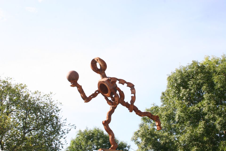 Skulptur einer rostigen Metallfigur, die einen Ball vor Himmel und Bäumen wirft.