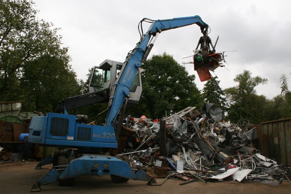 Blauer Bagger hebt Altmetall von einem Stapel auf einem Recyclinghof.
