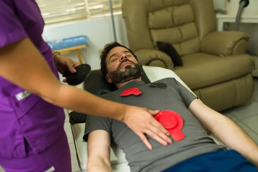 Persona que recibe tratamiento con dispositivos rojos en el pecho; trabajador de la salud en morado.