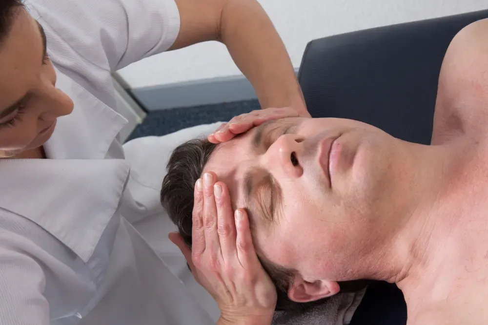 Un hombre recibe un masaje facial. Una persona con camisa blanca le aplica presión en la frente.