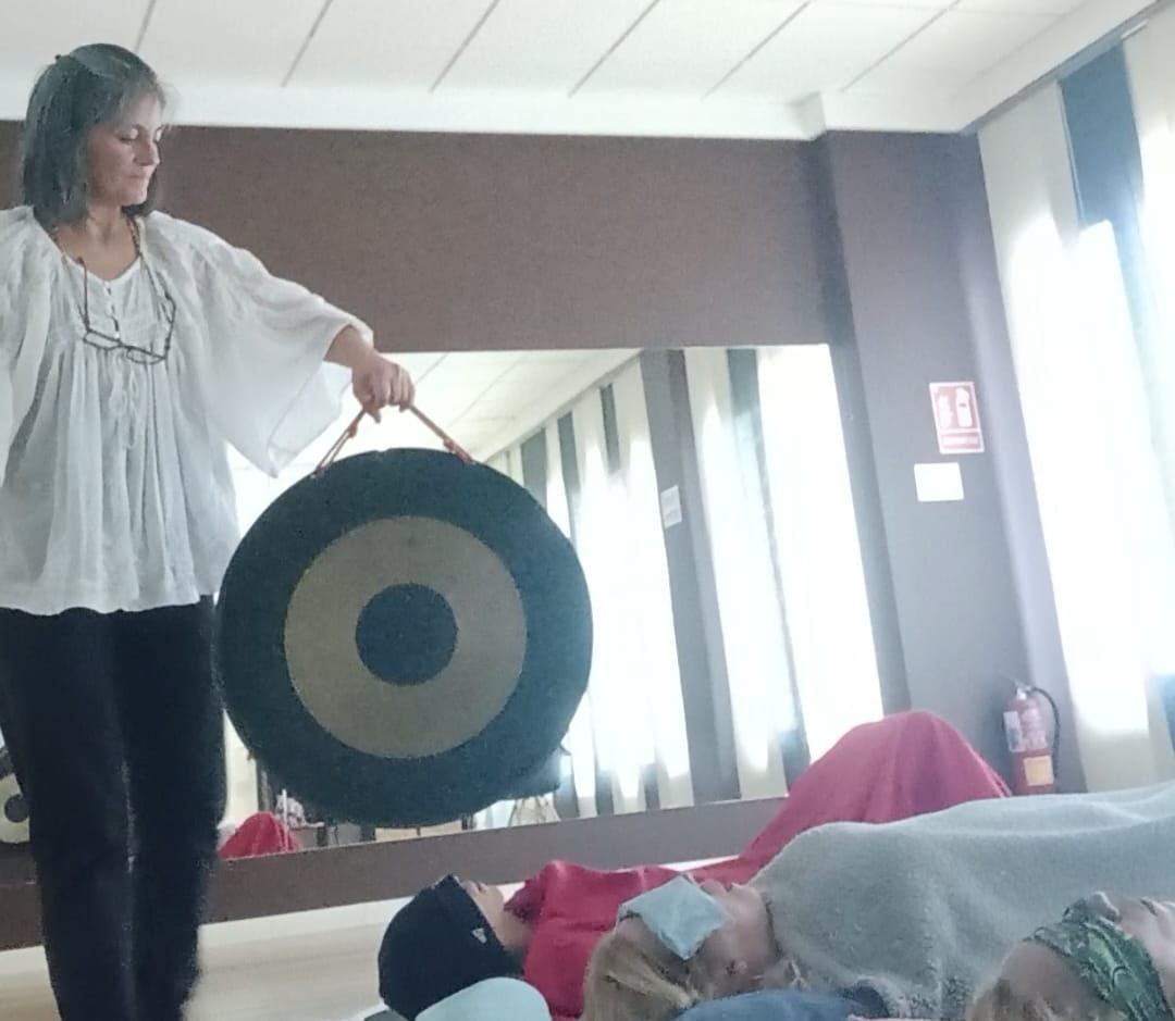 Mujer sosteniendo un gong, dirigiendo un baño de sonido para personas acostadas en una habitación.