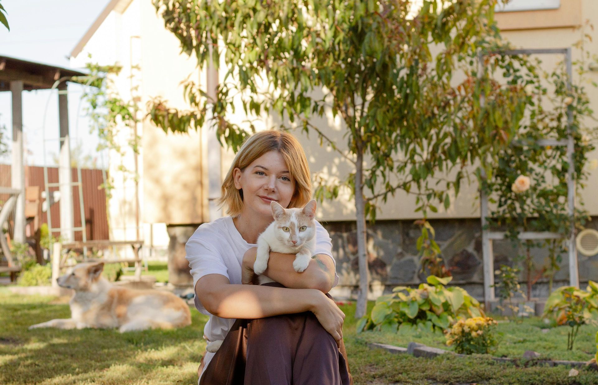 Una mujer con una camisa blanca sostiene un gato color crema y sonríe en un jardín iluminado por el sol; el perro es visible.