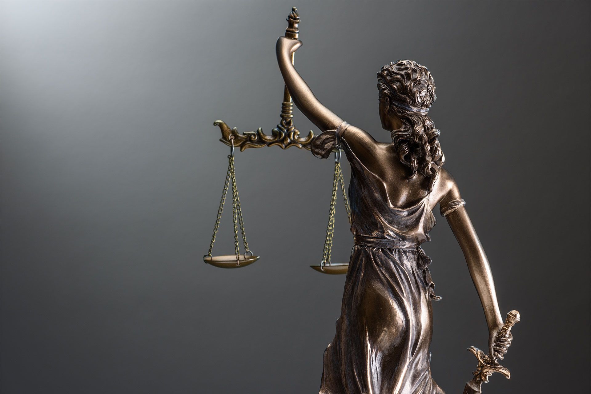 Statue de la Dame Justice tenant une balance et une épée, de dos, en bronze sur fond gris.