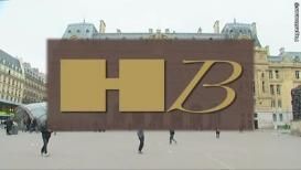 Panneau marron avec logo "HB" en doré, devant un bâtiment européen avec des personnes au premier plan.