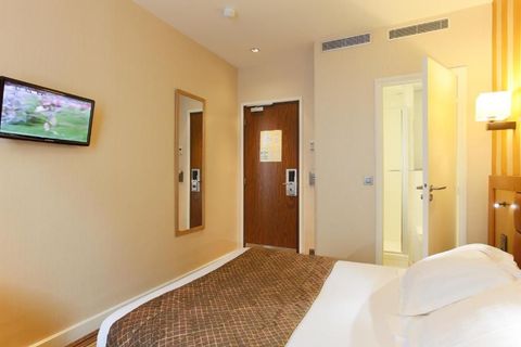 Intérieur d'une chambre d'hôtel avec lit, télévision, miroir et accès à la salle de bain. Murs beige clair.