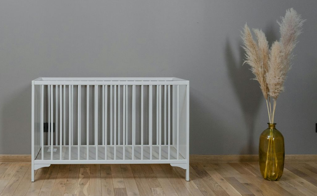 Berceau blanc contre un mur gris, à côté d'un vase d'herbe de la pampa sur un plancher en bois.