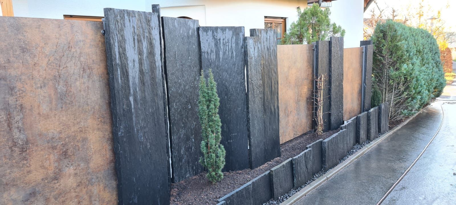 Ein moderner Sichtschutzzaun aus abwechselnd dunklen Steinen und braunen Paneelen mit einer kleinen Hecke.
