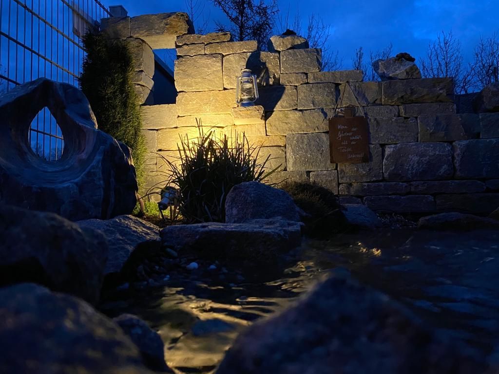 Steinmauer mit einem kleinen Teich, der in der Dämmerung von einem Scheinwerfer beleuchtet wird.