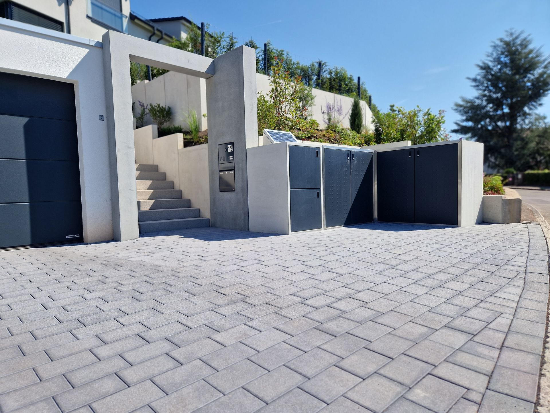 Eine graue Steinauffahrt führt zu einem modernen Gebäude mit dunklem Garagentor, Treppe und Mülltonnen. Sonniger Tag.