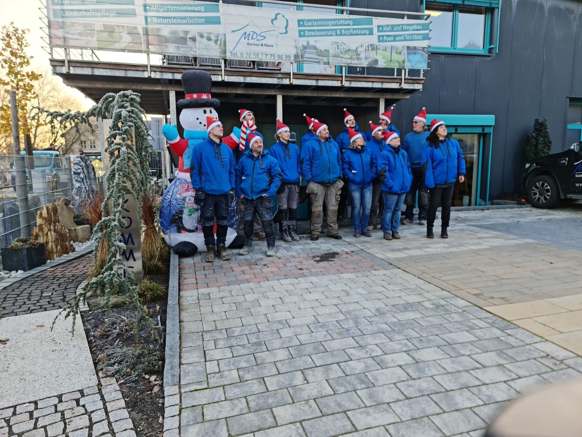 Eine Gruppe von Menschen in blauen Jacken und Weihnachtsmannmützen steht vor einem Gebäude, das mit einem Schneemann dekoriert ist.