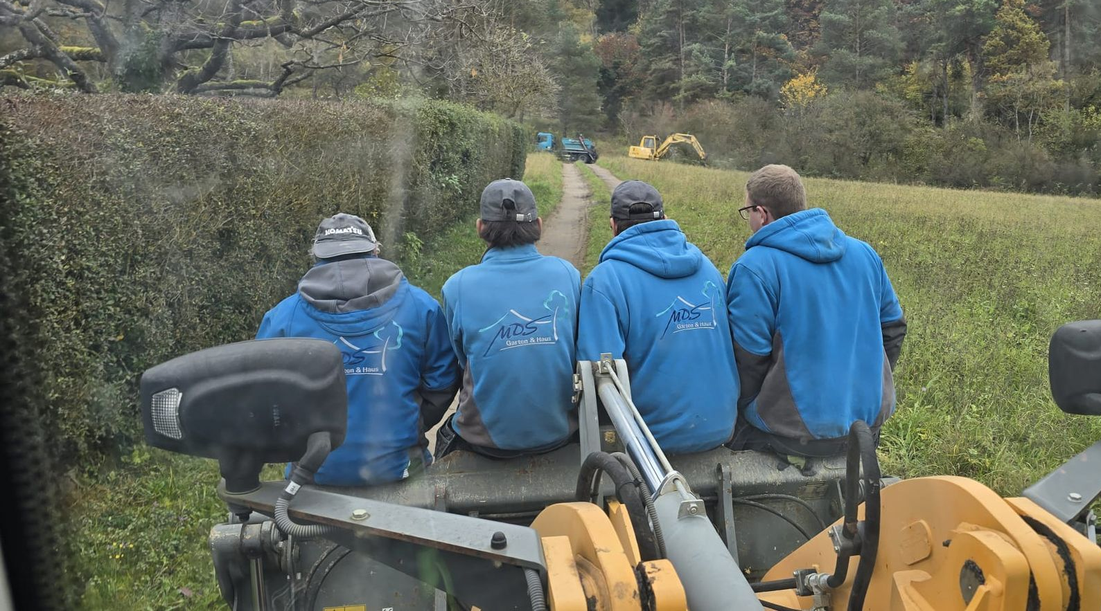 Vier Personen in blauen Jacken sitzen auf einem gelben Traktor, der auf einem grasbewachsenen Weg in ländlicher Umgebung fährt.