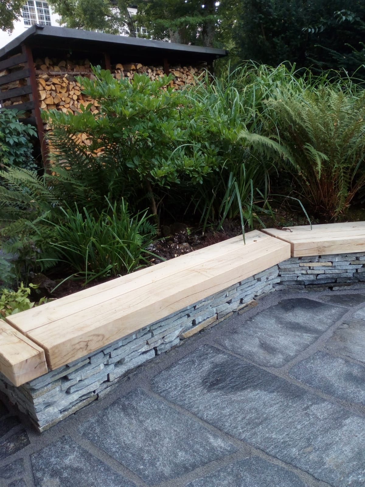Steinterrasse mit Steinmauer und Holzbank, im Hintergrund Grünpflanzen und ein Holzstapel.