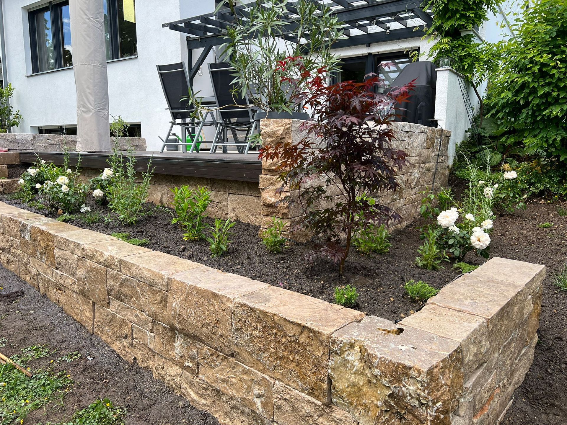 Ein Garten mit einer Steinmauer und einer Pergola vor einem Haus.