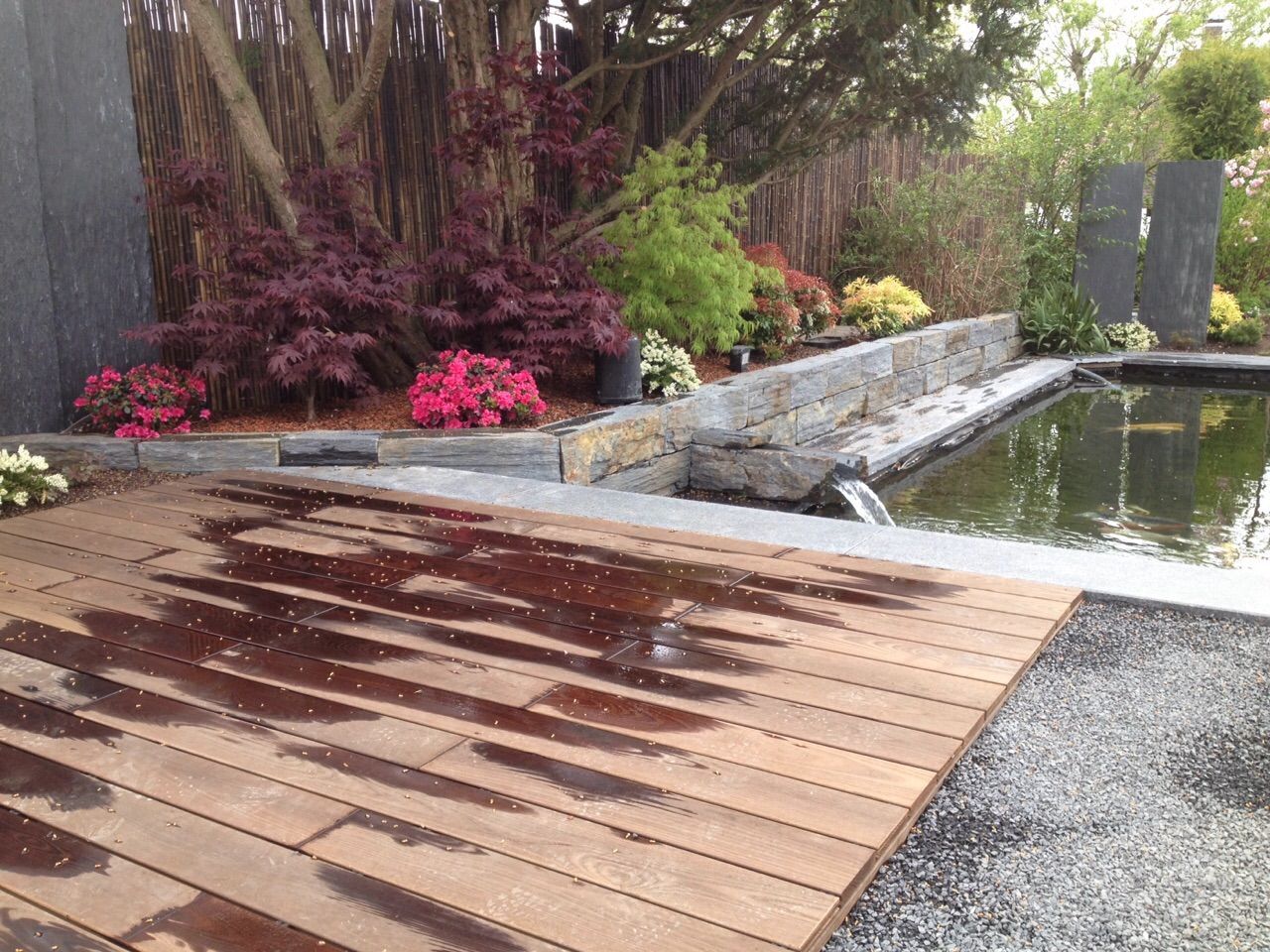Holzterrasse mit Wassertropfen, Blick auf einen Teich, Garten mit roten und grünen Pflanzen.