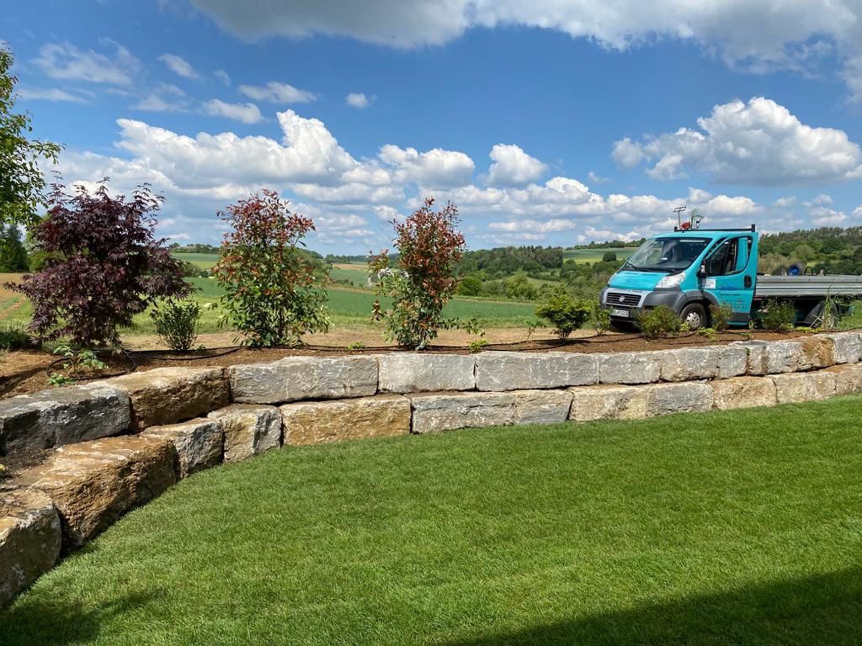 Ein blauer Lieferwagen steht auf einer Wiese neben einer Steinmauer.