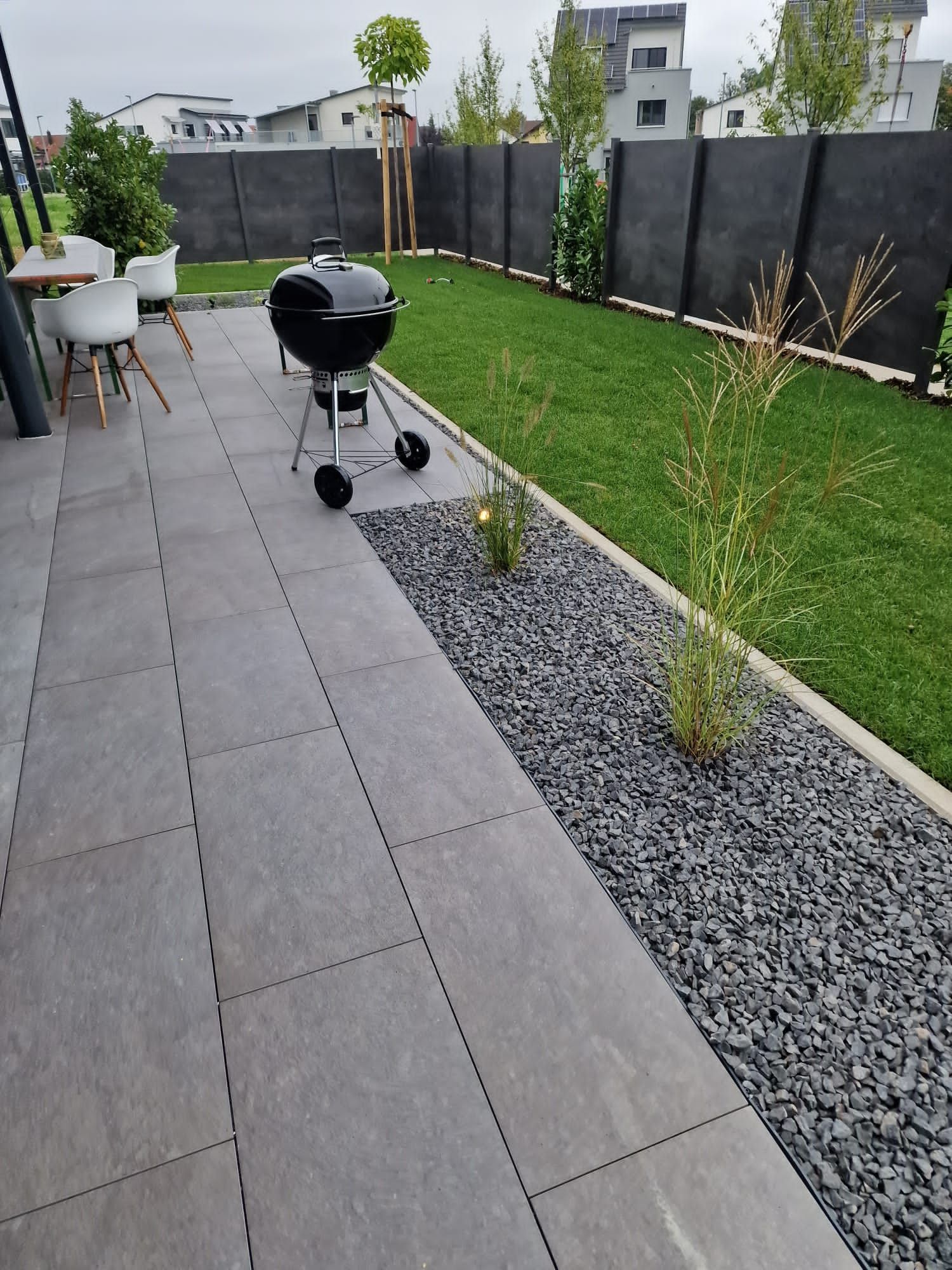 Terrasse mit grauen Steinfliesen, Grill, Rasen, schwarzem Kiesrand und schwarzem Zaun.