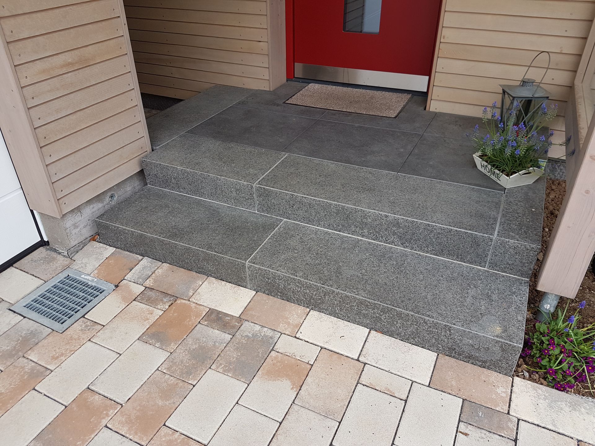 Steinstufen führen zu einer roten Tür mit einer Fußmatte; daneben stehen ein Blumentopf und eine Laterne.