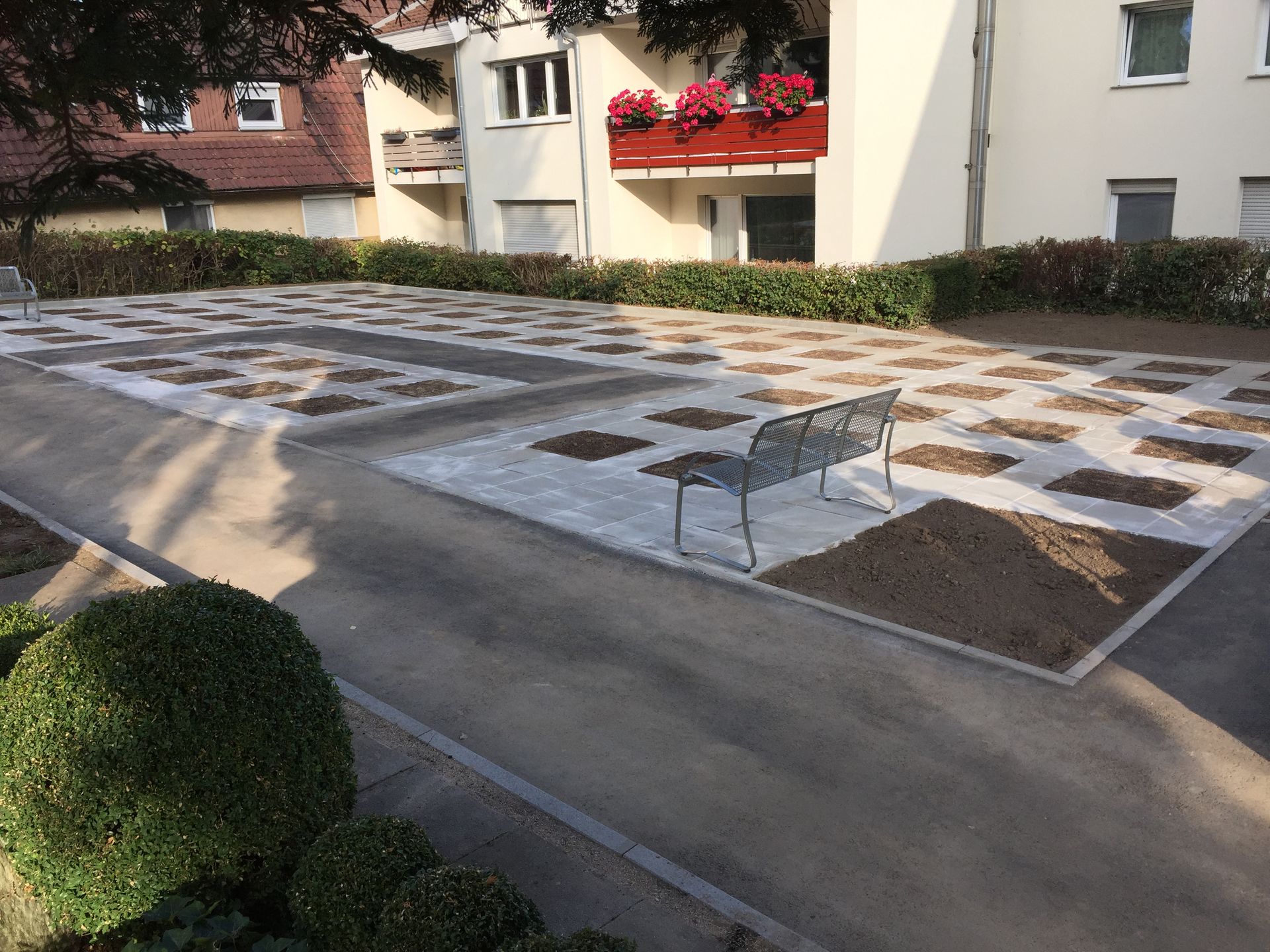 Eine Parkanlage mit Pflastersteinen, unbewachsenem Boden, einer Bank und umlaufenden Hecken. Im Hintergrund sind Gebäude zu sehen.