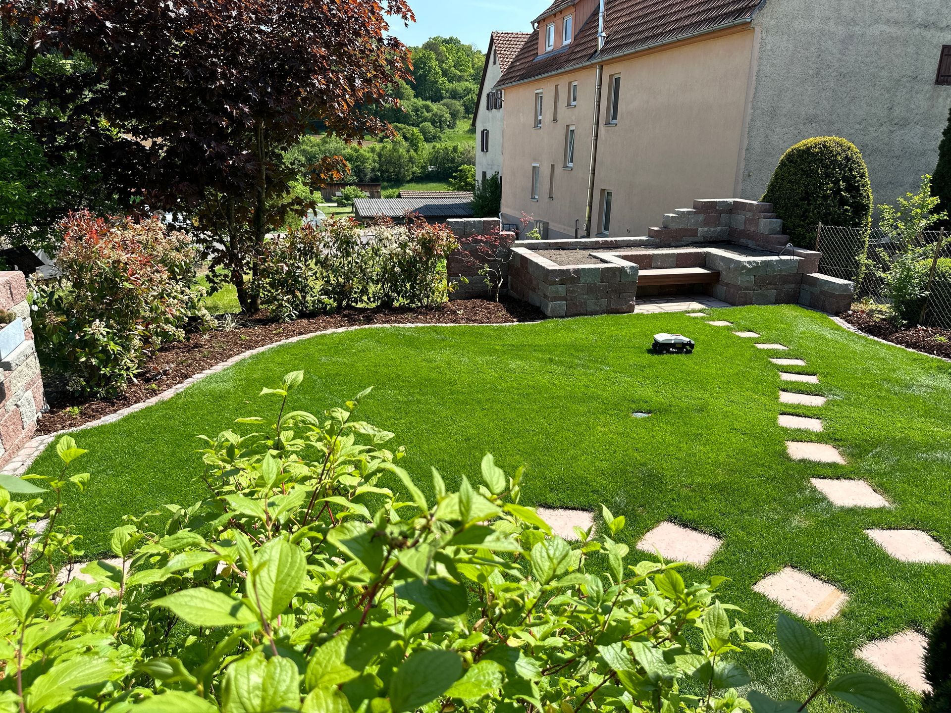 Grüner Rasen mit Trittsteinen, umgeben von Büschen und einer Steinmauer. Gebäude im Hintergrund.