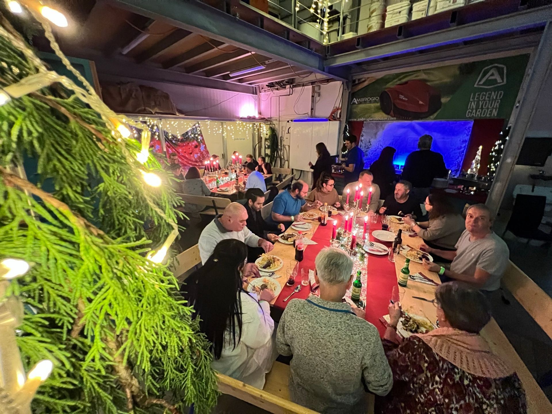 Menschen versammeln sich zu einem festlichen Abendessen an einem mit Lichtern und Grünpflanzen geschmückten Tisch in einem Innenraum.