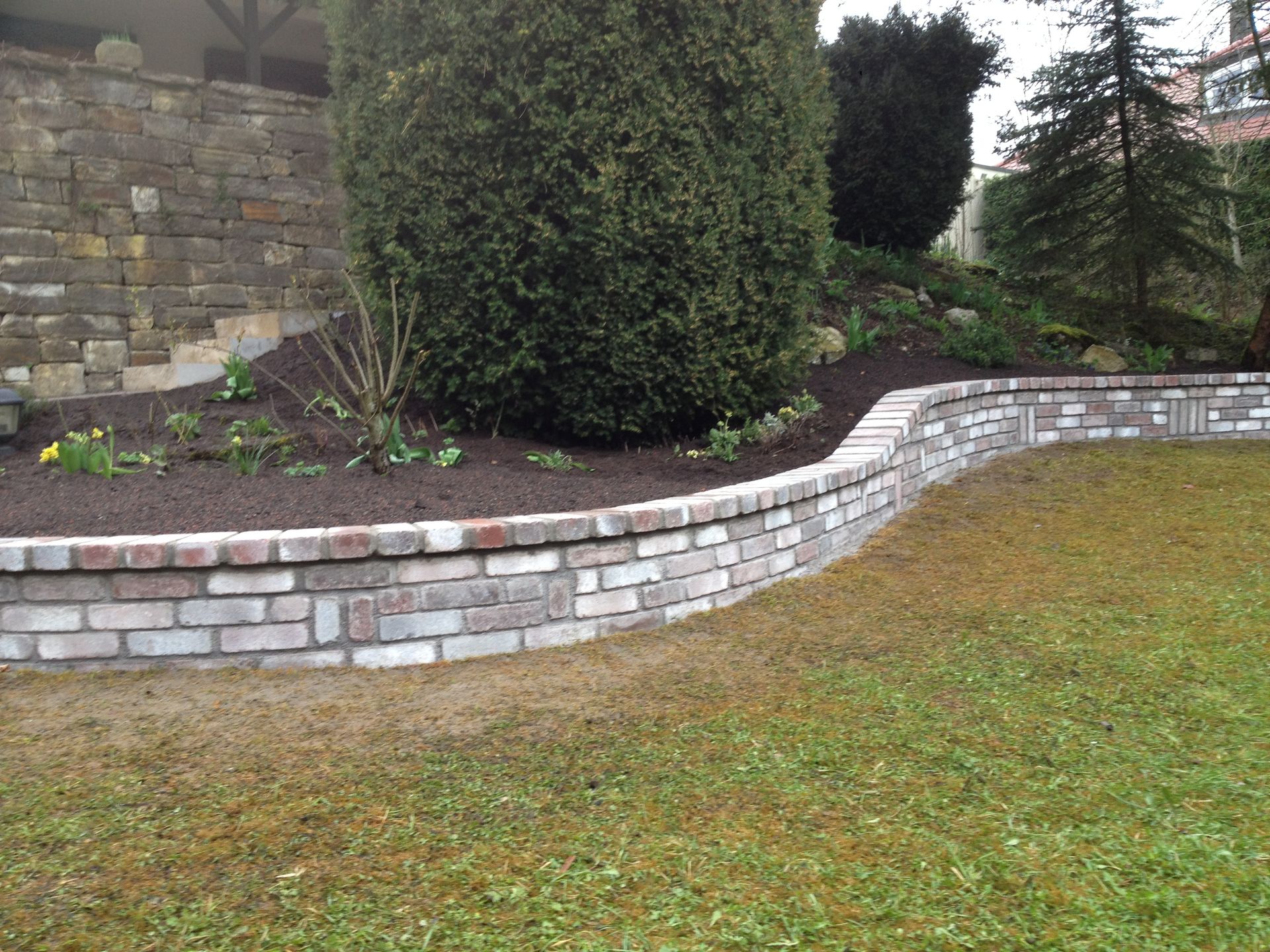 Eine Stützmauer aus Ziegelsteinen mit einem Beet aus dunklem Mulch, grünen Pflanzen und Rasen.