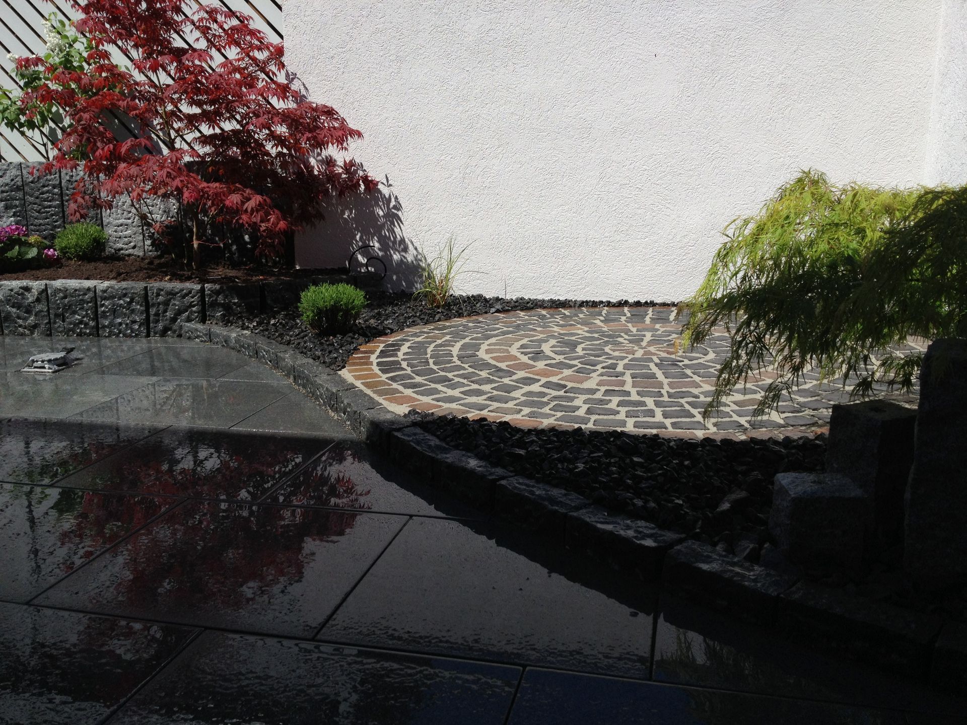Garten mit einer kreisförmigen, mit Kopfsteinpflaster belegten Terrasse, die von dunklem Gestein und Pflanzen umgeben ist, mit einem roten Ahorn und einem hängenden grünen Baum vor einer weißen Mauer.