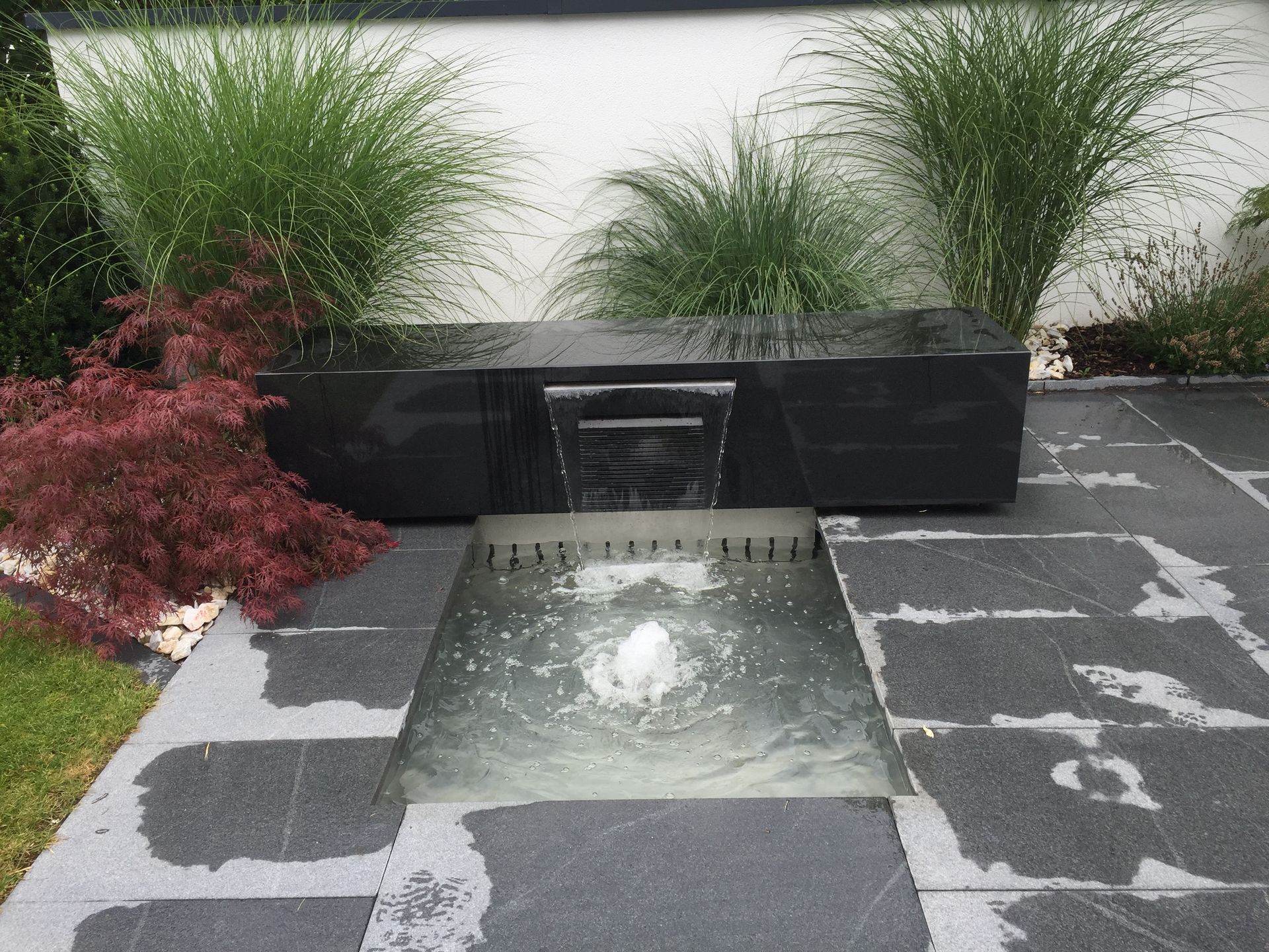 Ein Springbrunnen mit schwarzem Steinelement, aus dem Wasser in ein Becken kaskadenartig herabfließt. Im Hintergrund Ziergräser und rotes Laub.
