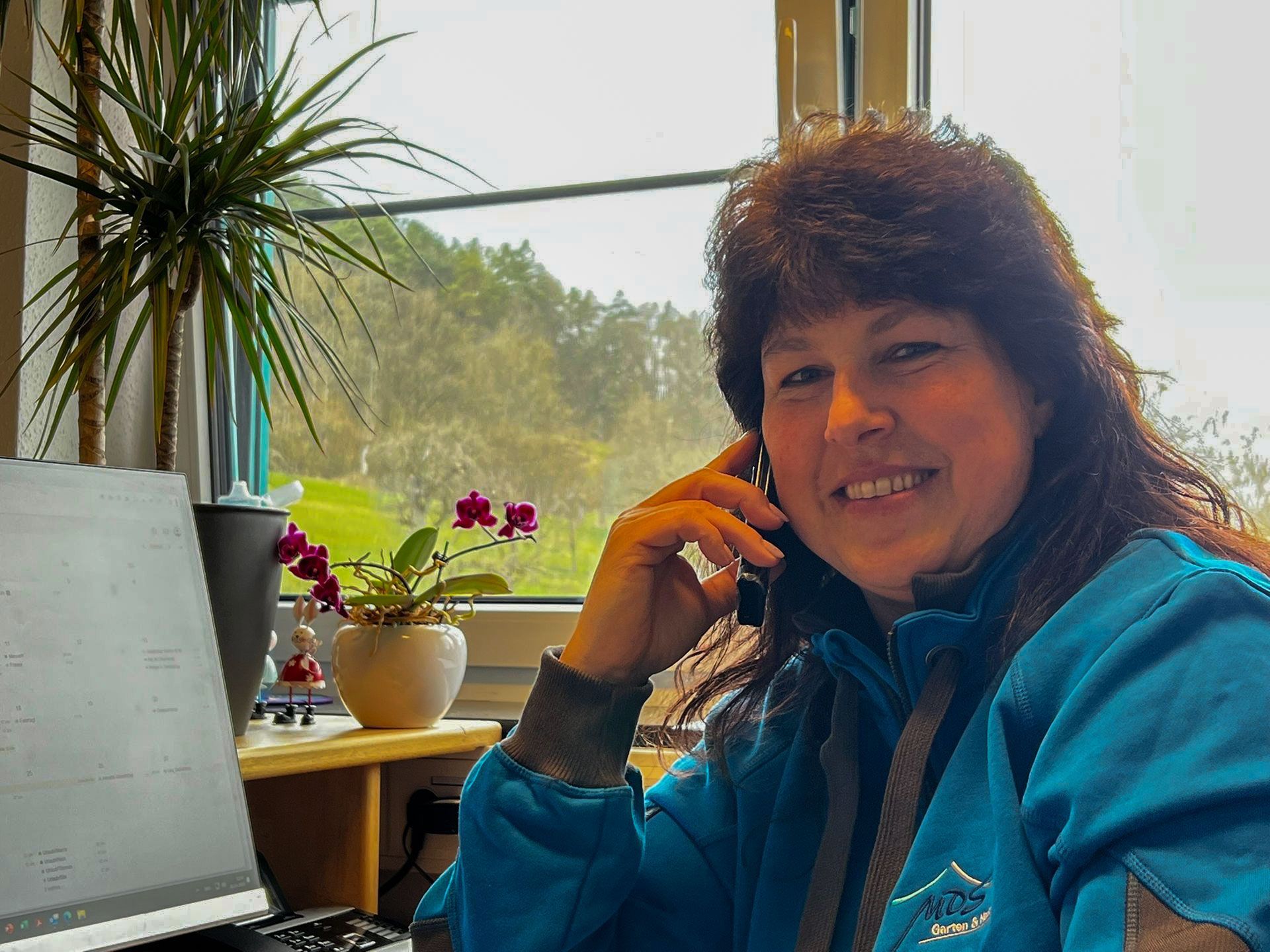 Eine Frau in blauer Jacke lächelt während eines Telefonats in der Nähe eines Fensters und von Pflanzen.