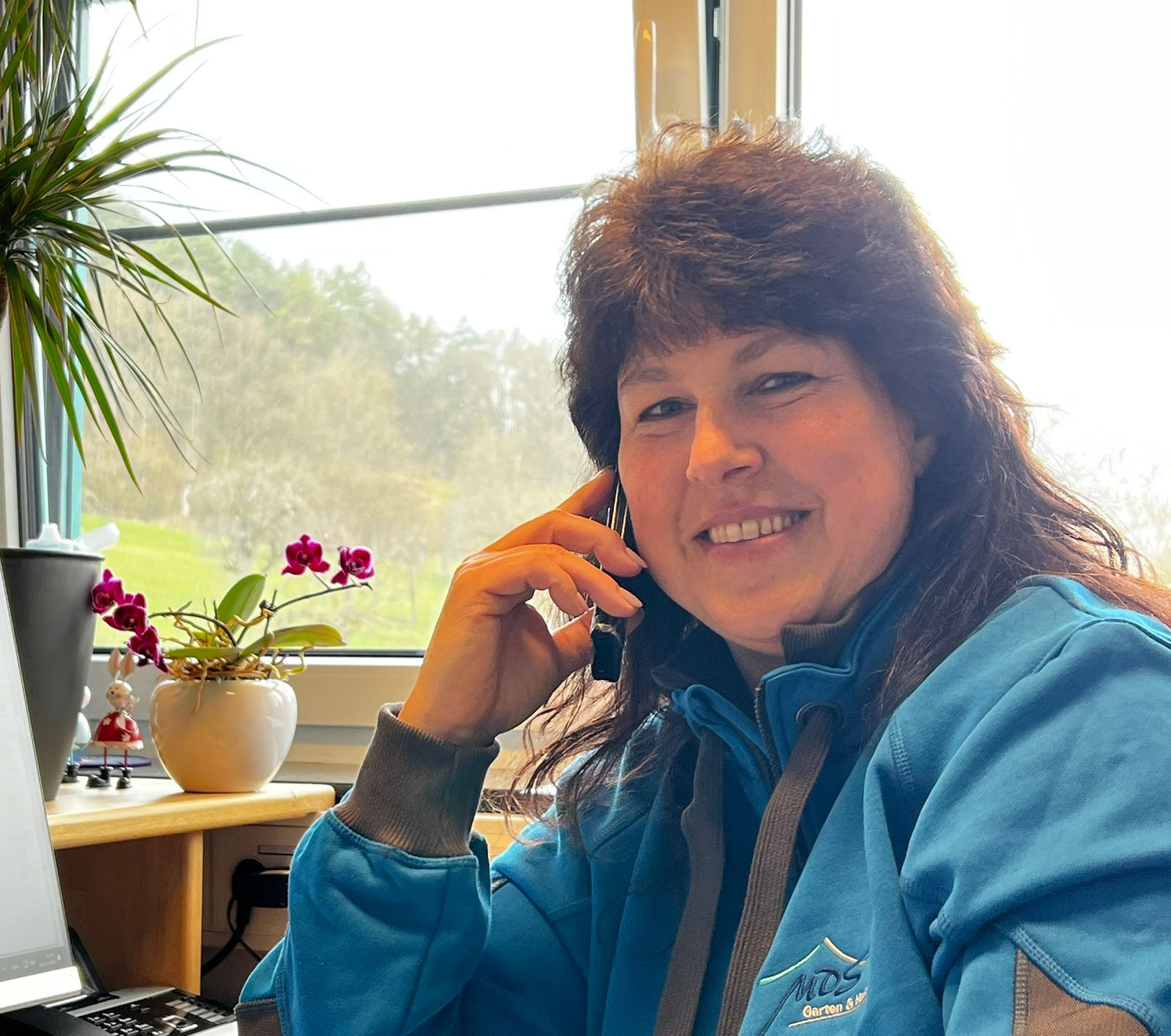 Frau in blauer Jacke telefoniert, lächelt am Fenster, Büroumgebung mit Pflanzen.