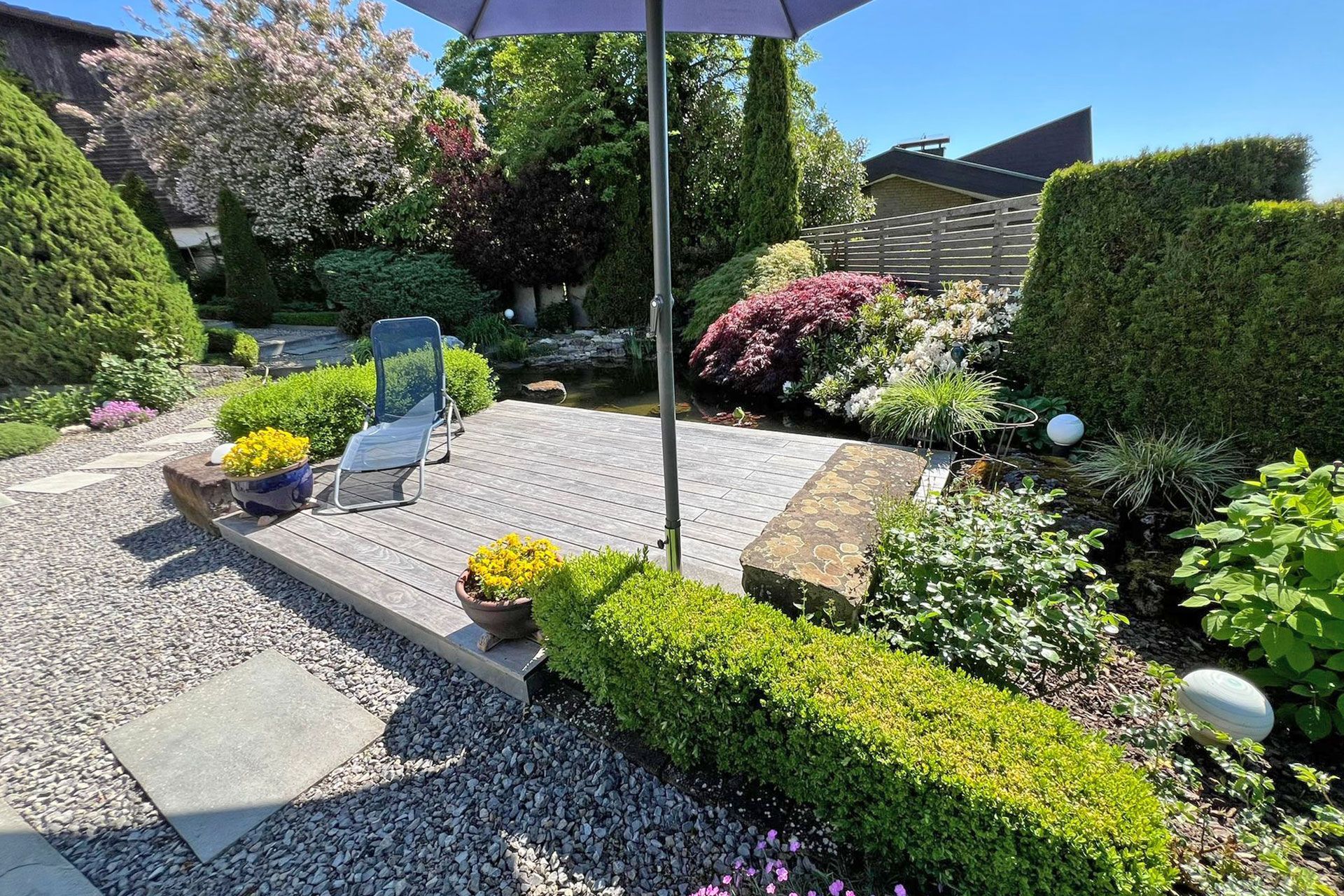 Terrasse mit Kies, akkurat geschnittenen Hecken, Sonnenschirm, Stuhl und kleinem Teich. Sonnige Lage im Freien.
