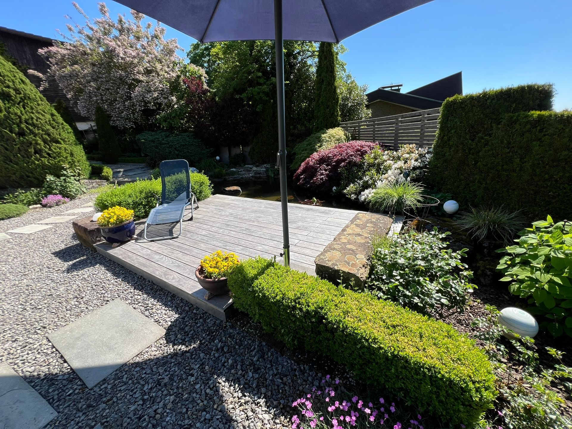 Terrasse mit Kies, akkurat geschnittenen Hecken, Sonnenschirm, Stuhl und kleinem Teich. Sonnige Lage im Freien.