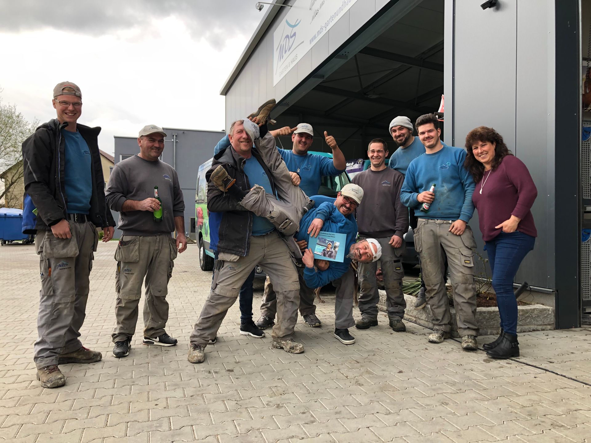 Eine Gruppe von Menschen posiert im Freien, einige lächeln. Ein Mann wird kopfüber gehalten. Graue und blaue Kleidung.