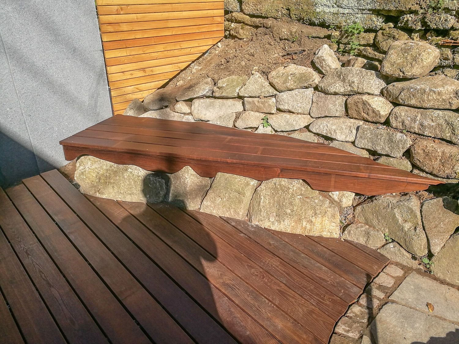 Eine Holzbank steht auf einem Holzdeck neben einer Steinmauer.