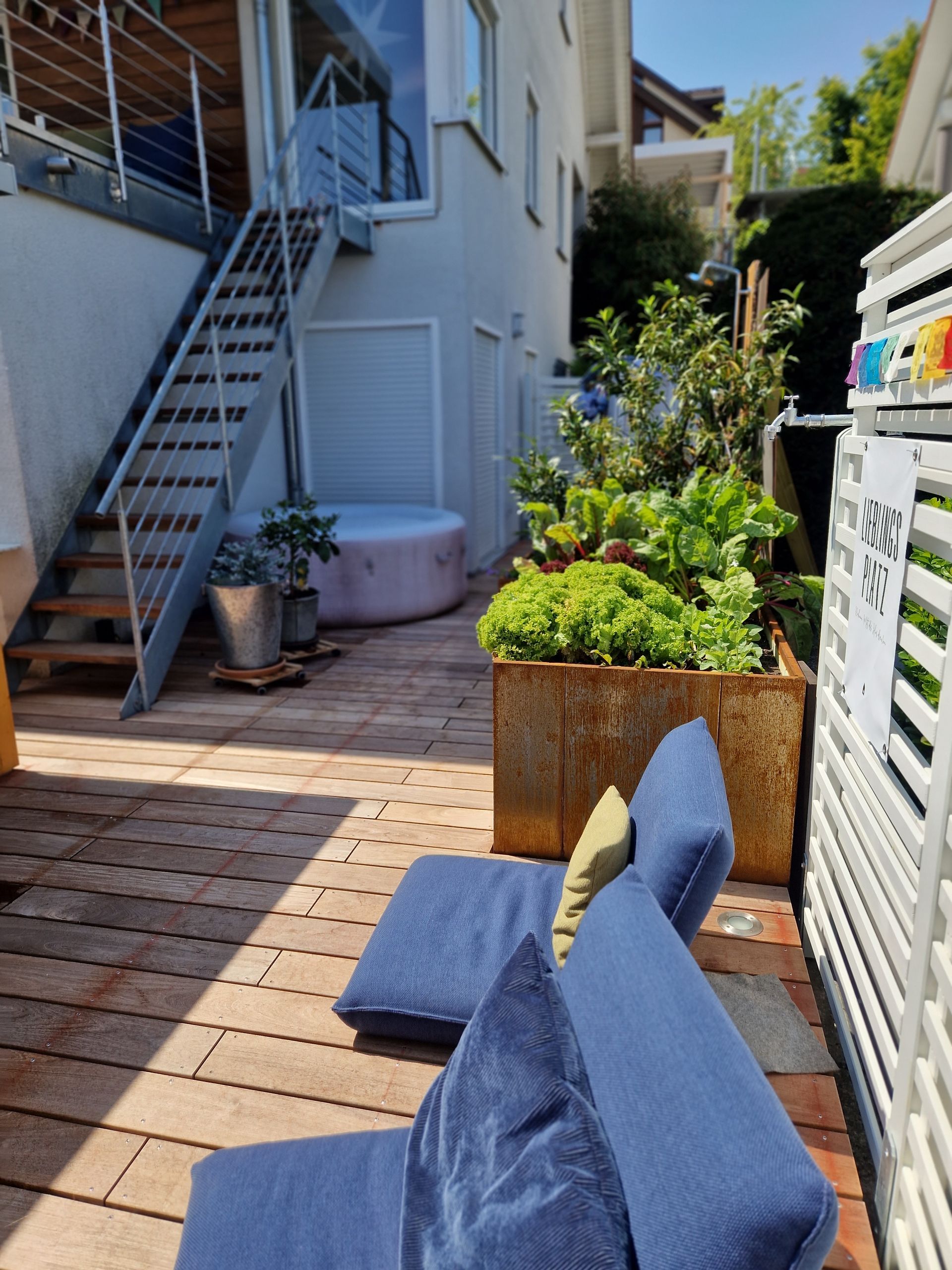Blauer Liegestuhl auf einer Holzterrasse; Topfpflanzen, Metalltreppe und ein Whirlpool sind zu sehen.